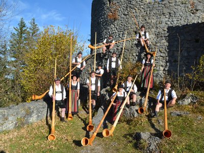 Zehn Personen in traditioneller Tracht stehen und sitzen um eine alte Steinmauer. Sie halten große Alphörner. Die Männer tragen schwarze Hüte und weiße Hemden, die Frauen bunte Röcke.