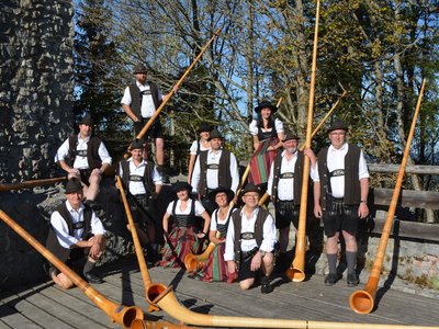 Gruppe von zwölf Personen in traditioneller Tracht, posierend mit mehreren Alphörnern. Einige tragen schwarze Westen und weiße Hemden, andere haben farbige Röcke.