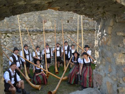 Gruppe von zwölf Personen in traditioneller Tracht, die große Holzblasinstrumente halten. Sie stehen in einer steinernen Ruine mit Bögen.